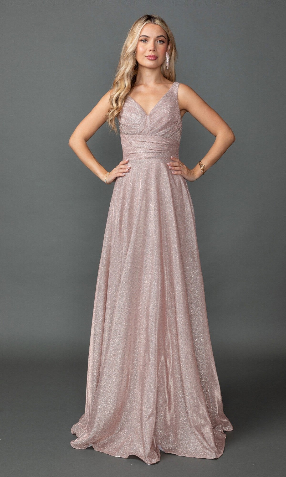 A woman poses against a gray backdrop wearing Dancing Queen 4545—a long, elegant prom dress in light pink with a sleeveless design, V-neckline, fitted waist, and sparkling glitter details.